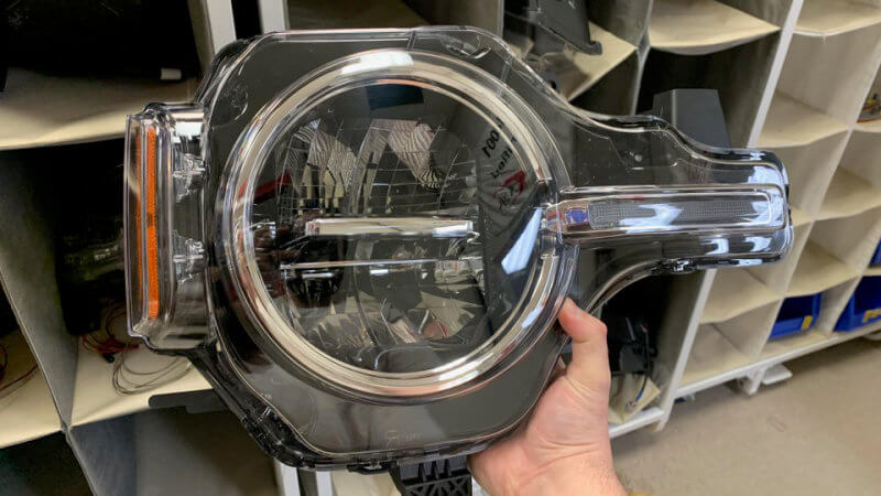 Mão de uma pessoa segura peça do farol redondo do Ford Bronco com modura cromada, seta laranja integrada e seção lateral de LED que deve se integrar à grade