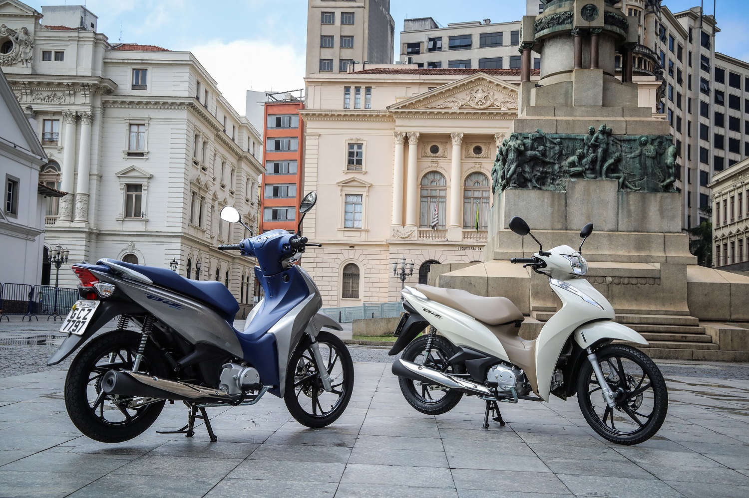 Honda Biz 2020 de traseira na cor prata com banco azul ao lado branca de frente de uma