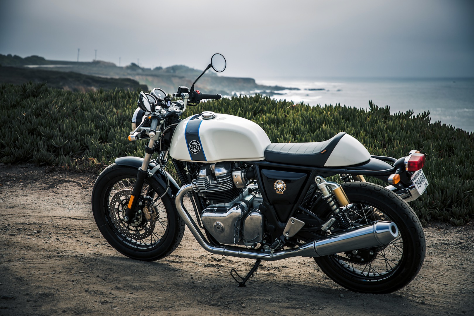 Royal Enfield Continental GT estática de traseira com tanque branco com faixa esverdeada e tendo o mar ao fundo