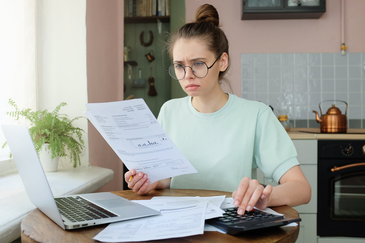 Imagem mostra mulher de óculos e com cabelo preso com papel na mão, calculadora na mesa e à frente de um notebook