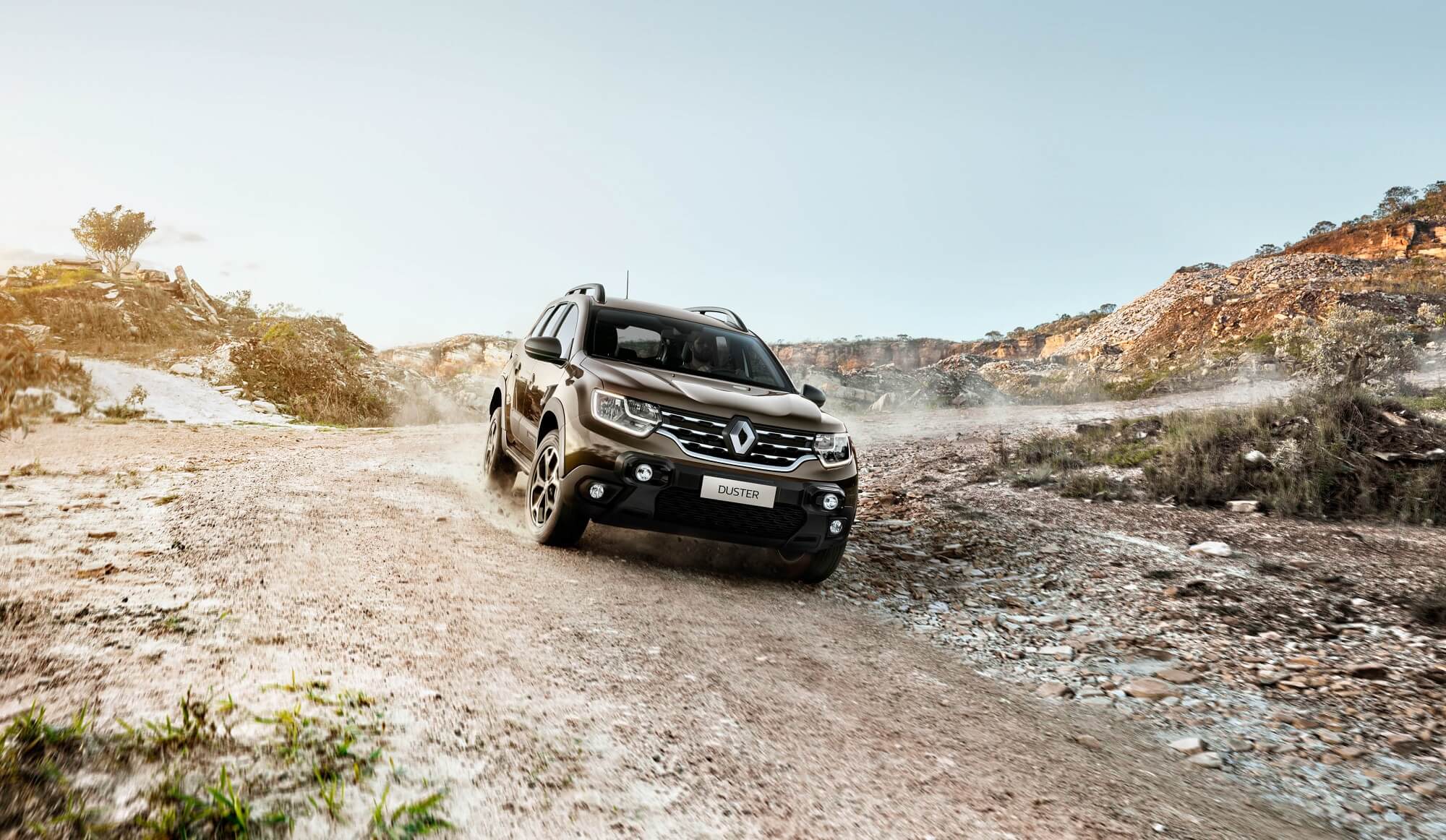 Novo Renault Duster marrom metálico m vovimento de frente em estrada de terra e pedrinhas com penhasco árido ao fundo