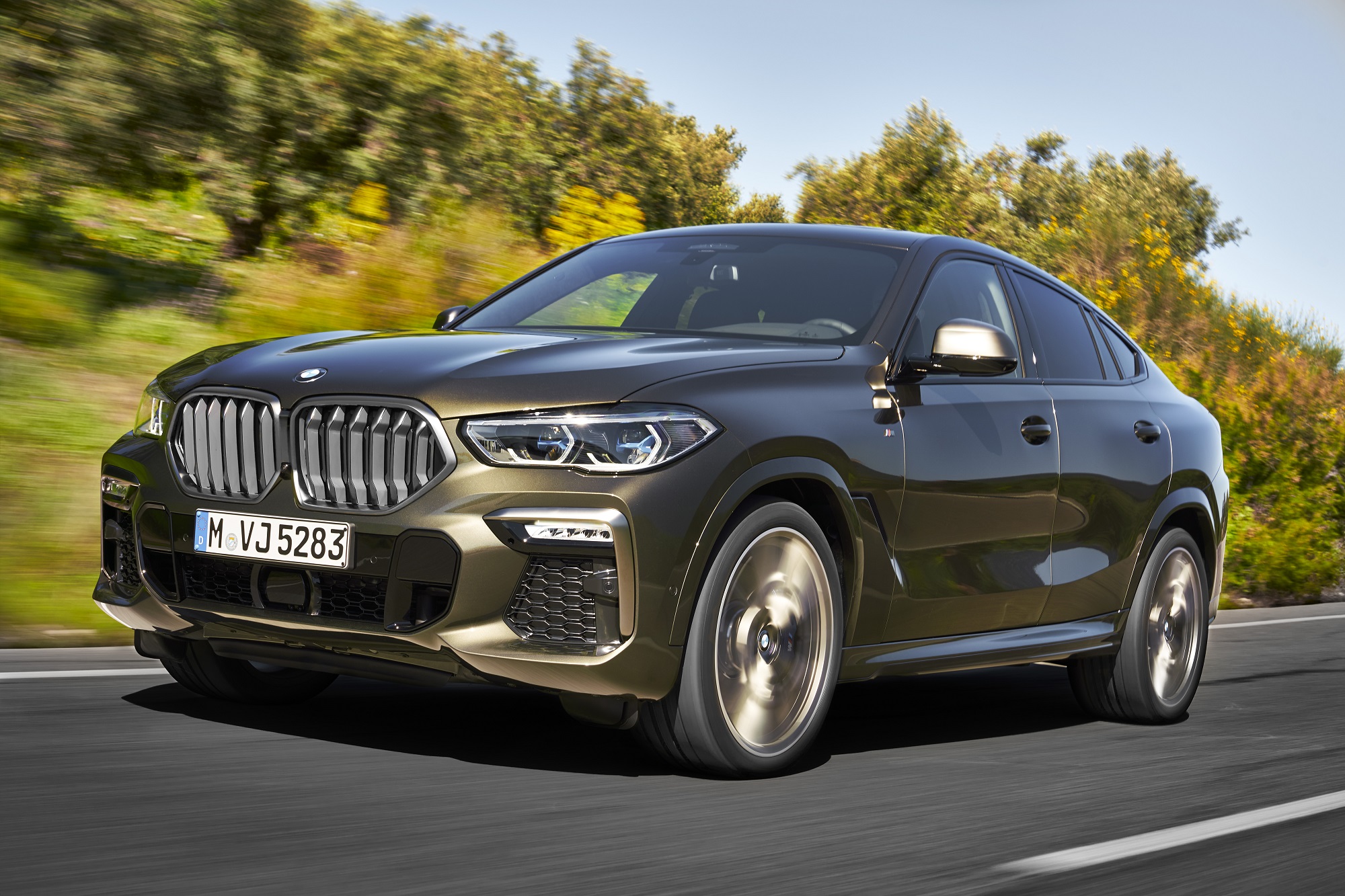 Bmw X6 marrom metálico em movimento na estrada com enorme grade dianteira e árvores ao fundo