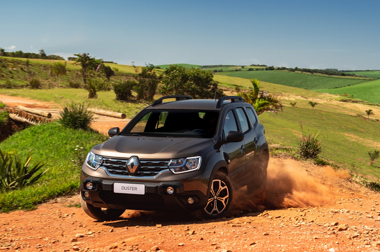 Novo Renault Duster em movimento fazendo curva para a esquerda em estrada de terra levatnadndo um pouco de poeira e um externso gramado e colinas ao fundo