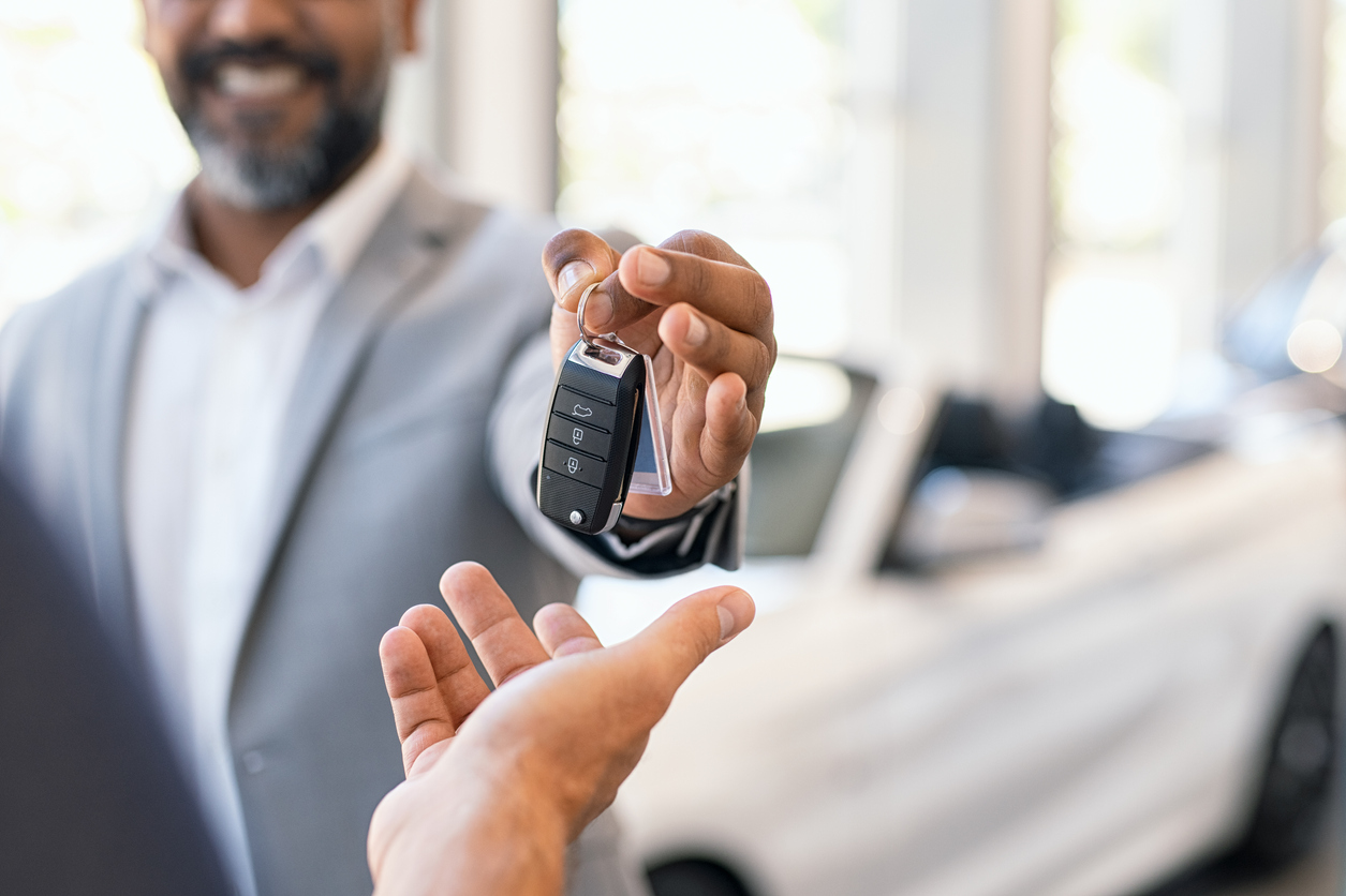 Vendedor que não tem o rosto totalmente revelado segura chave de carro e entrega na mão de uma outra pessoa em uma concessionária de veículos
