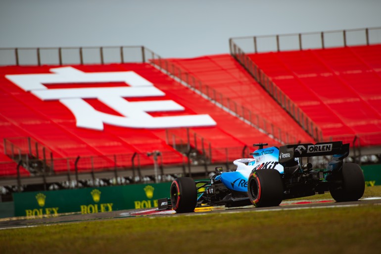 Carro de Fórmula 1 de traseira na tomada de curva em treino e ao fundo a arquibancada vermelha do GP de Pequim