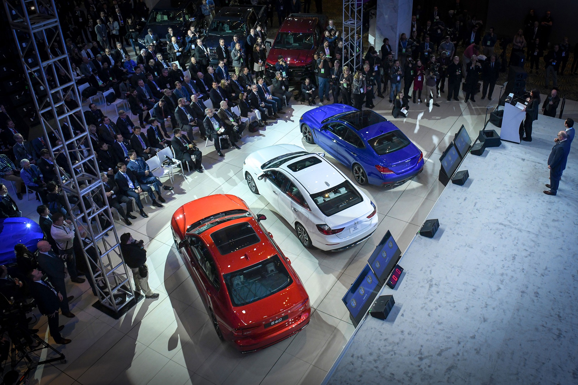 Imagem de cima mostra coletiva de imprensa do Salão De Detroit 2019 com apresentador em um palco ao lado de três caros e vários jornalistas acompanhando