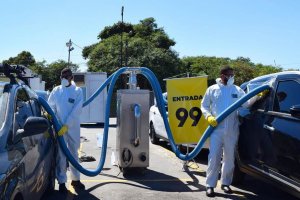 Dois funcionários com roupa anti-contaminação branca e máscaras seguram as mangueiras da tecnologia de limpeza para higienizar os carros no estacionamento em Guarulhos