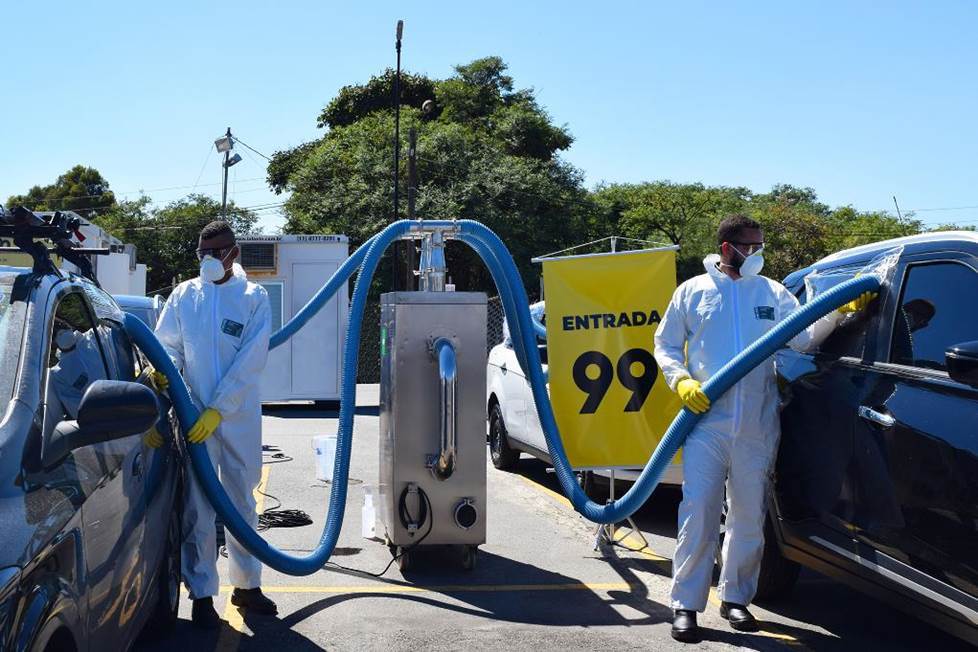 Dois funcionários com roupa anti-contaminação branca e máscaras seguram as mangueiras da tecnologia de limpeza para higienizar os carros no estacionamento em Guarulhos