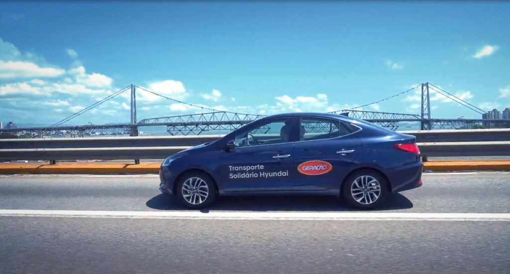 Hyundai HB20S em movimento na estrada com adesivos do programa Transporte Solidário e tendo ao fundo a ponte Hercílio Luz, em Florianópolis