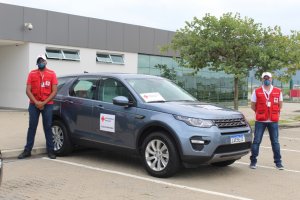 Dois funcionários com máscaras de proteção ao lado de um Land Rover Discovery com adesivos da Cruz Vermelha Brasileira