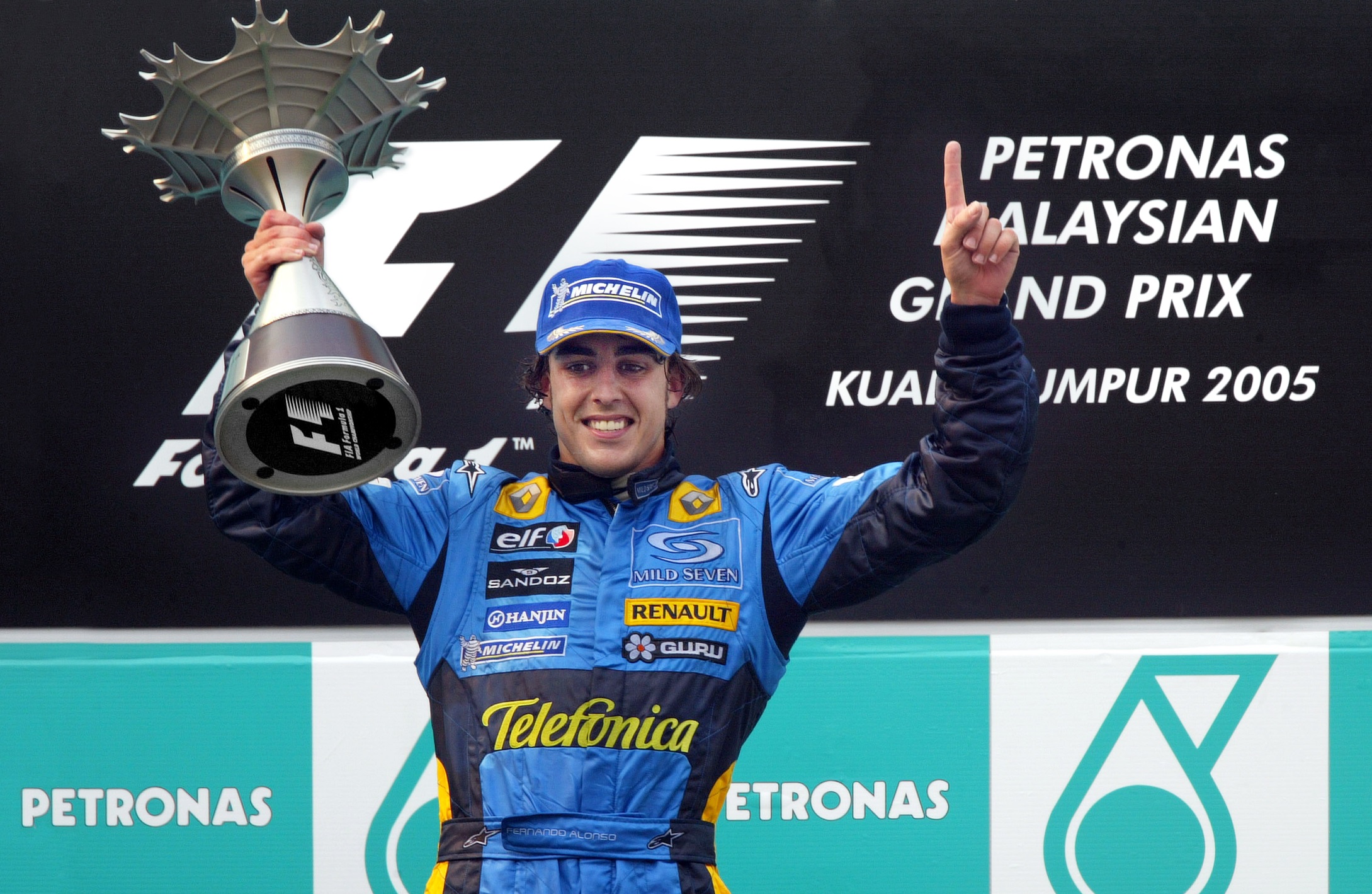 Fernando Alonso segura a taça no pódio e com a outra mão faz o 1 de primeiro