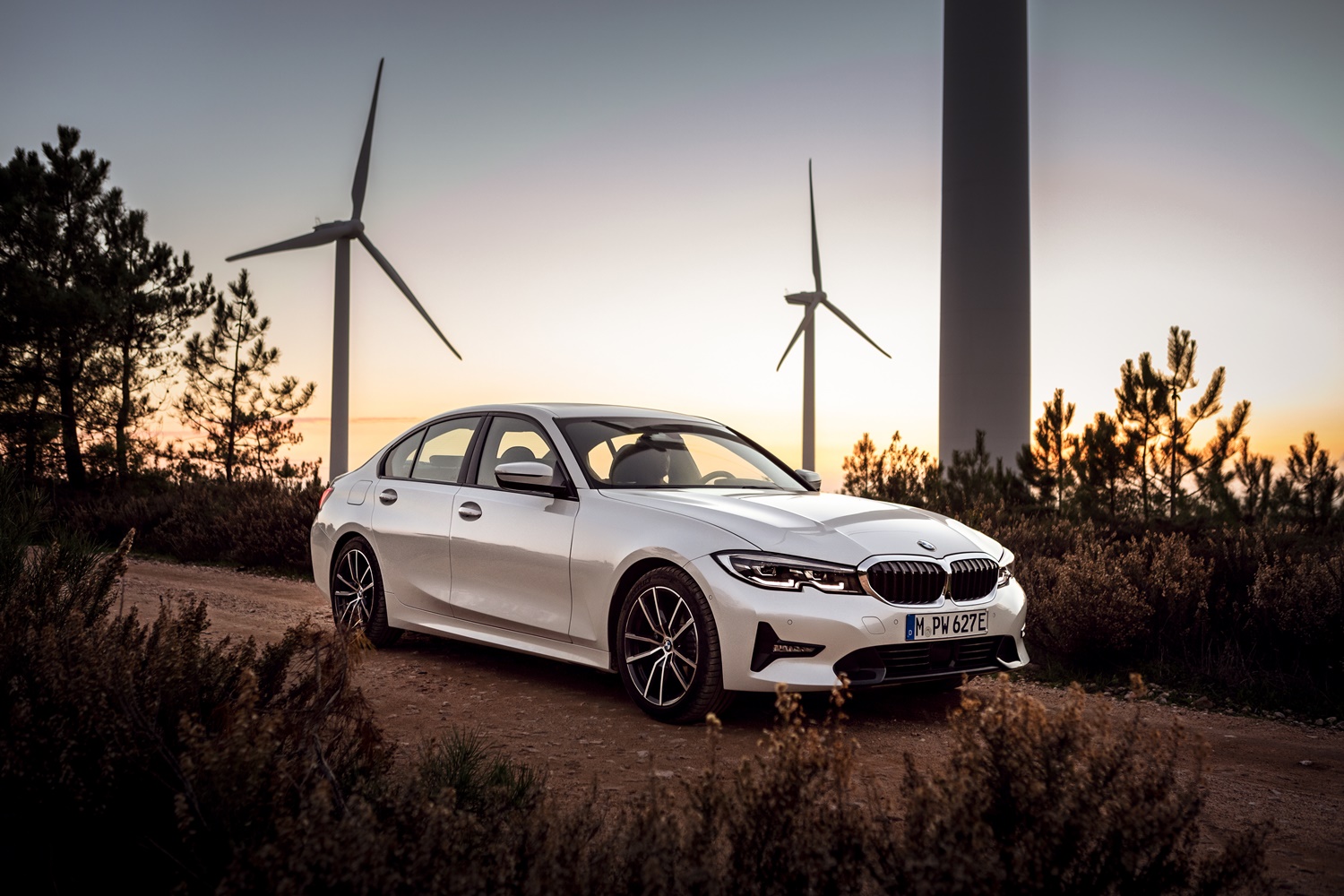 Bmw Série 3 branco em uma estrada com moinhos de energia eólica ao fundo no entardecer