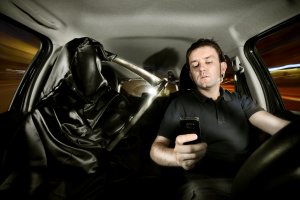 Imagem mostra um motorista escrevendo uma mensagem de texto no celular enquanto dirige e ao seu lado uma pessoa vestida de morte