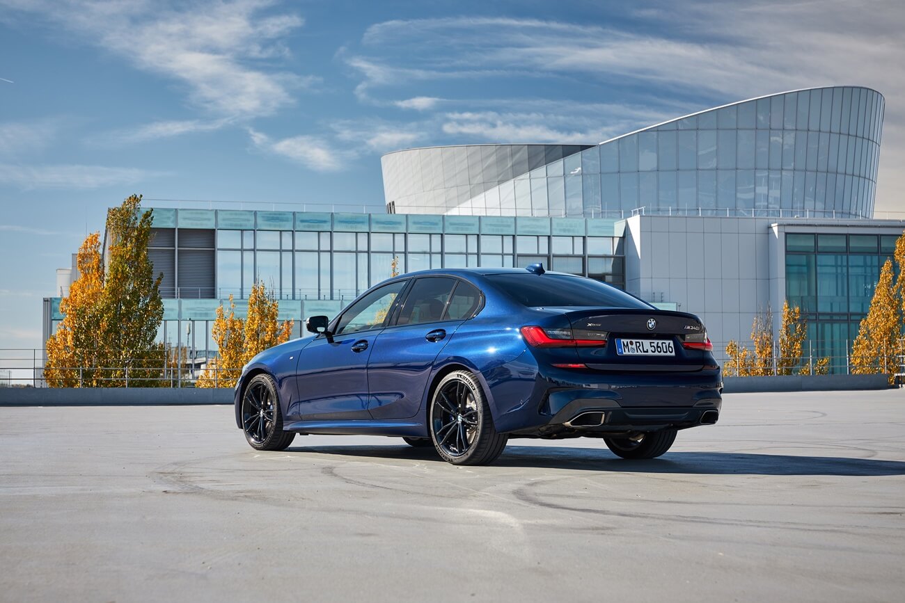 BMW M340i xDrive azul de traseira com prédio baixo espelhado ao fundo