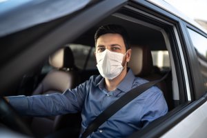 motorista homem, vestindo uma camisa azul e com cinto de segurança, digirindo com máscara de proteção