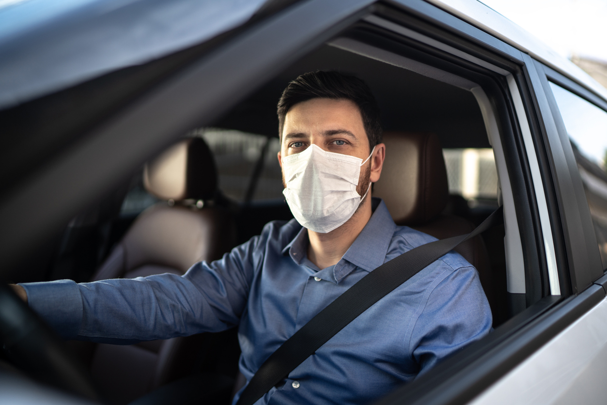 motorista homem, vestindo uma camisa azul e com cinto de segurança, digirindo com máscara de proteção